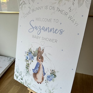 May include: A white baby shower sign with a watercolor design of leaves and flowers. The sign says "Some bunny is on their way! Welcome to Suzanne's Baby Shower". A cartoon bunny rabbit is in the bottom center of the sign.