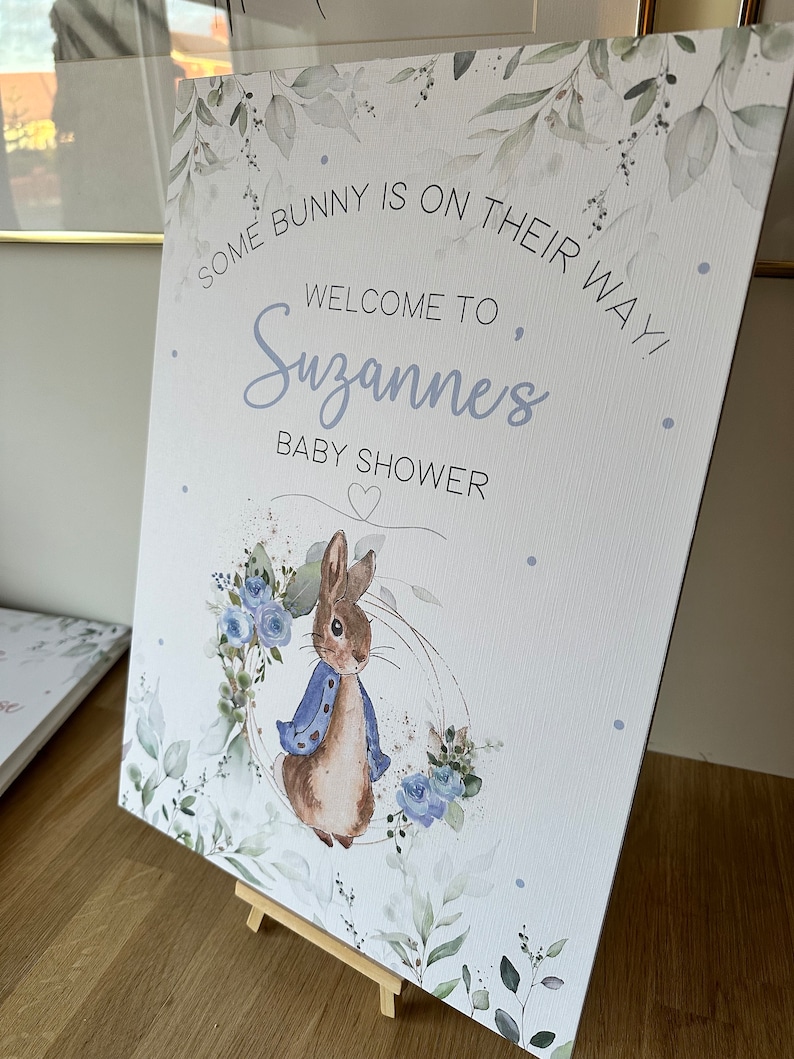 May include: A white baby shower sign with a watercolor design of leaves and flowers. The sign says "Some bunny is on their way! Welcome to Suzanne's Baby Shower". A cartoon bunny rabbit is in the bottom center of the sign.