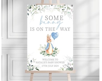 May include: A baby shower welcome sign on a wooden easel. The sign features a watercolor illustration of a bunny holding a blue balloon, with the text "Some bunny is on the way". Decorative greenery surrounds the text and image.