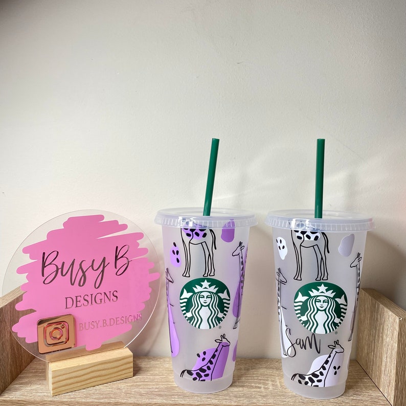 May include: Two clear plastic Starbucks tumblers with green straws. The tumblers feature a giraffe design in black and purple. The Starbucks logo is visible on the front of the tumblers. The text "Sam" is written on one of the tumblers.