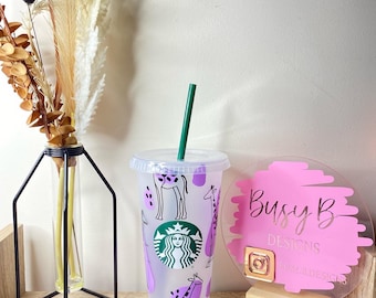May include: A clear plastic tumbler with a green straw, featuring a giraffe pattern in black and purple. The Starbucks logo is visible. A pink sign with gold text and a wooden base is to the right.