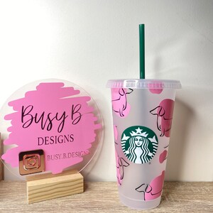 May include: A clear plastic cold cup with a green straw and lid. The cup features a pink dachshund pattern and a Starbucks logo. A pink and clear sign with the text "Busy B DESIGNS" is to the left.