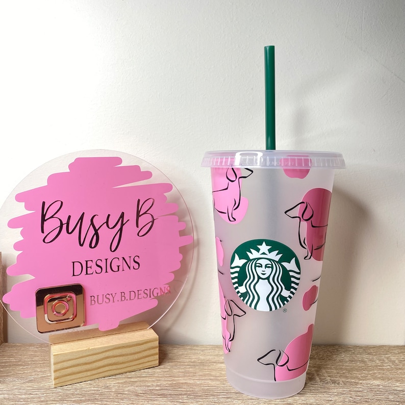 May include: A clear plastic cold cup with a green straw and lid. The cup features a pink dachshund pattern and a Starbucks logo. A pink and clear sign with the text "Busy B DESIGNS" is to the left.