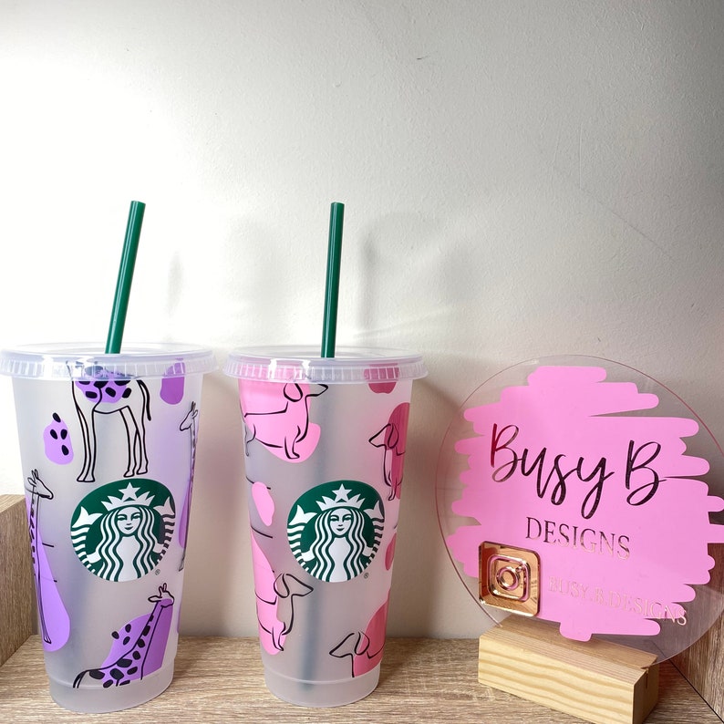 May include: Two clear plastic tumblers with green straws. One features a purple giraffe pattern, the other a pink dachshund pattern. Both have the Starbucks logo. A pink sign with the text "Busy B Designs" is also visible.