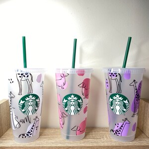 May include: Three clear plastic Starbucks tumblers with green straws. The tumblers feature animal designs: giraffes and dogs in pink and purple. One tumbler has the name "Sam" written on it. The Starbucks logo is visible on each cup.