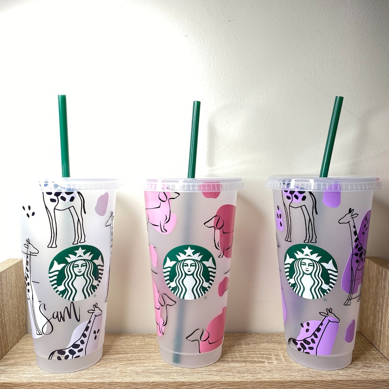 May include: Three clear plastic Starbucks tumblers with green straws. The tumblers feature animal designs: giraffes and dogs in pink and purple. One tumbler has the name "Sam" written on it. The Starbucks logo is visible on each cup.