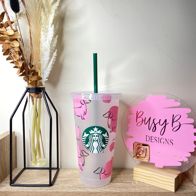 May include: A clear plastic tumbler with a green straw, featuring a pink dachshund pattern and the Starbucks logo. The tumbler is displayed on a wooden shelf with a decorative vase and a pink sign that reads "Busy B Designs".
