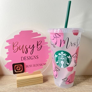 May include: A clear plastic Starbucks tumbler with a green straw, featuring pink dachshund dog and spot designs. The tumbler has the name "Mrs D" in black script. A pink and gold "Busy B Designs" sign is to the left.