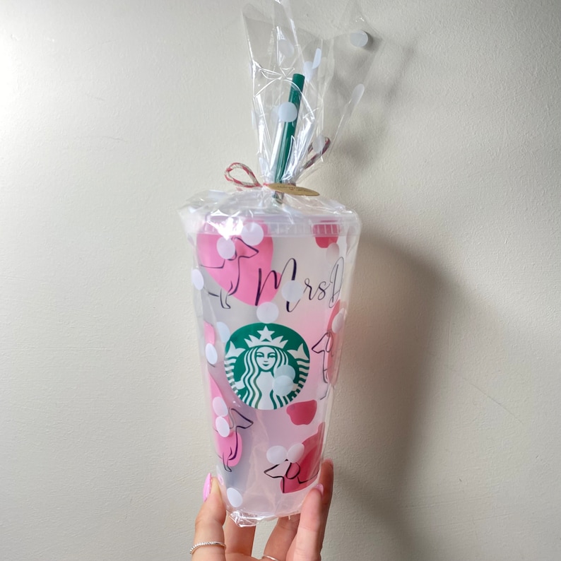 May include: Clear plastic Starbucks tumbler with pink dachshund and heart designs, white polka dots, and the word "Mrs" in script. The tumbler has a green straw and is wrapped in cellophane.