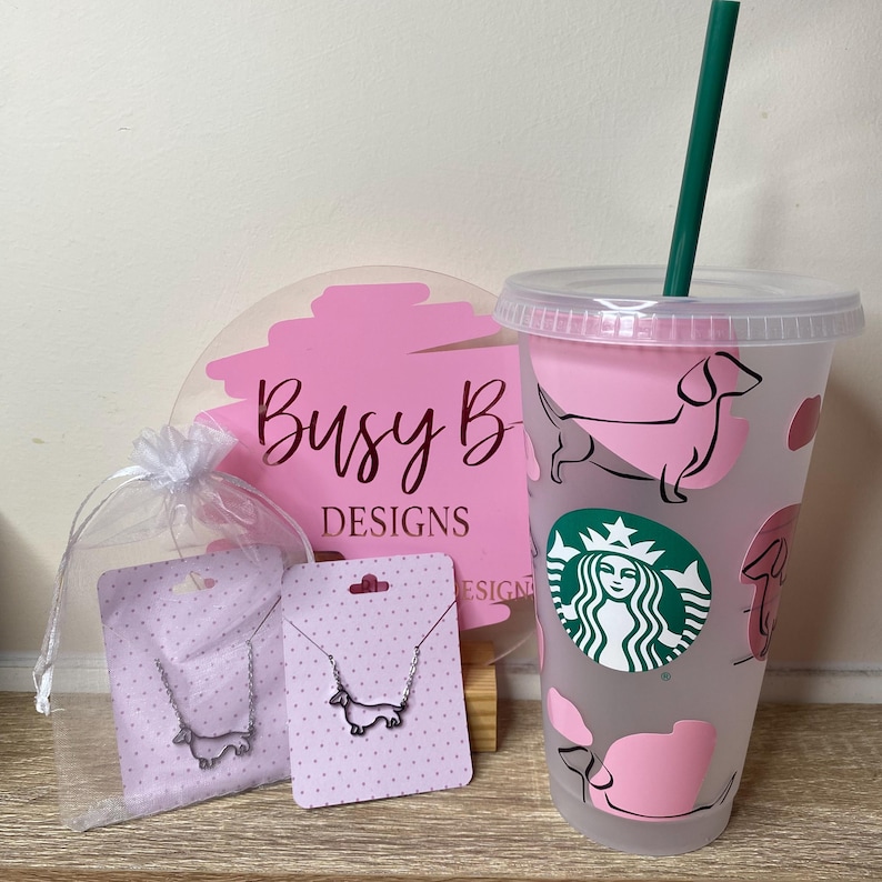 May include: A clear plastic Starbucks tumbler with a pink dachshund design and a green straw. A pink sign with the text "Busy B DESIGNS" is behind the tumbler. Two dachshund necklaces are displayed on pink cards.