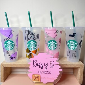 May include: Four clear plastic Starbucks tumblers with green straws. Each tumbler features a different design: giraffes, the name "Miss Nonkett", pink dachshunds, and a dachshund with the name "Ruth". A pink sign with "Busy B Designs" is in front.
