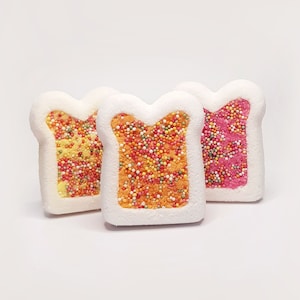 May include: Three toast-shaped bath bombs with white crusts and colourful centres. The left one has a yellow centre, the middle one is orange, and the right one is pink. Each centre is covered in small, multi-coloured round sprinkles.