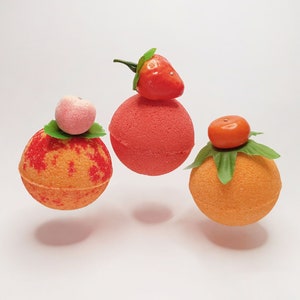 May include: Three spherical bath bombs in shades of red, orange, and peach. Each is adorned with a fruit topper: a strawberry, a peach, and an orange. The bath bombs are presented against a plain background, giving the impression they are floating.