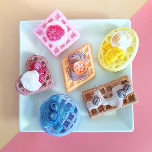 May include: A selection of decorative waffles in various shapes and colours, presented on a white plate. The waffles are adorned with toppings such as fruit and cream. The plate is set against a pink and yellow backdrop, creating a vibrant display.
