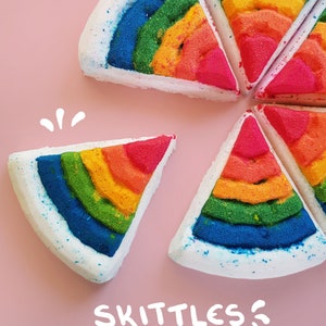 May include: Rainbow-coloured bath bombs in the shape of pizza slices. Each slice has layers of red, orange, yellow, green, and blue, with a white base and blue glitter. The word "SKITTLES" is written in white script on a pink background.