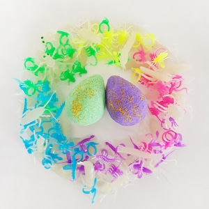 May include: Two egg-shaped bath bombs, one mint green and one purple, are surrounded by a rainbow of plastic insect toys. The bath bombs have gold glitter accents. The insects are in shades of white, yellow, green, blue, pink, and purple.