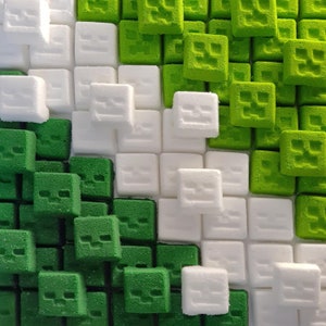 May include: A close-up of a pixelated design made of green and white square blocks. The green blocks feature a face design, while the white blocks are plain. The blocks are arranged in a grid pattern, creating a textured surface.
