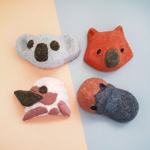 May include: Four animal-shaped bath bombs. A grey koala, an orange wombat, a pink and white platypus, and a brown and grey echidna. The bath bombs are on a peach and blue background.