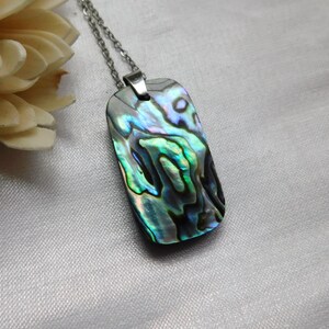 May include: A rectangular pendant necklace with a shimmering abalone shell design. The pendant features iridescent blues, greens, and purples, and is suspended from a silver-colored chain. The necklace is a piece of jewelry.