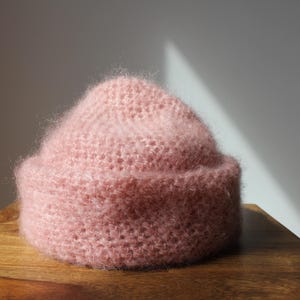 Könnte beinhalten: Eine hellrosa Mütze mit flauschiger, texturierter Oberfläche und einem breiten, umgeschlagenen Rand. Die Mütze ist in einem sichtbaren Strickmuster gefertigt und weist ein weiches, gebürstetes Erscheinungsbild über die gesamte Krone auf.