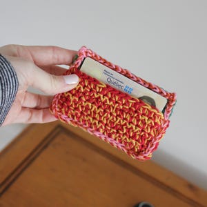 May include: A handmade crochet cardholder in red, yellow, and pink. The cardholder is rectangular with a textured stitch pattern. A card with the text "Québec" is partially visible inside the holder. The edges are finished with a pink and green border.