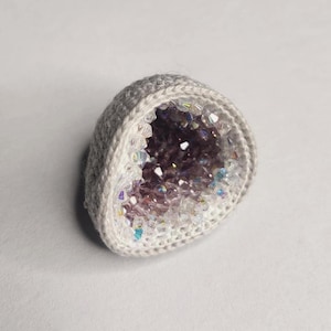 May include: A dome-shaped object featuring a light gray crocheted exterior and a hollow center filled with iridescent, multifaceted clear and light purple crystals that catch the light, creating a sparkling, textured appearance throughout the interior.