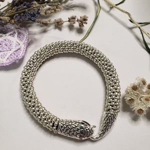 May include: A silver snake bracelet with a textured snake head clasp. The bracelet is made of small, round beads.