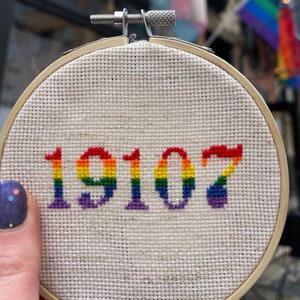 May include: A rainbow-colored cross-stitch embroidery hoop with the number 19107 stitched in the center.