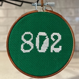 May include: A green embroidery hoop with a white cross-stitch design of the number 802.