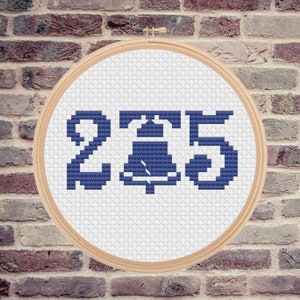 May include: A blue cross-stitch embroidery hoop with the number 2, a bell, and the number 5. The bell is in the center of the hoop.