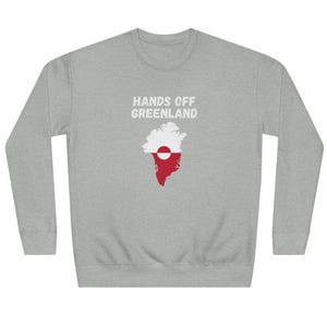 Op de afbeelding: Een lichtgrijs sweatshirt met de tekst "HANDS OFF GREENLAND" boven een afbeelding van Groenland in rood en wit. De sweatshirt heeft een ronde hals.