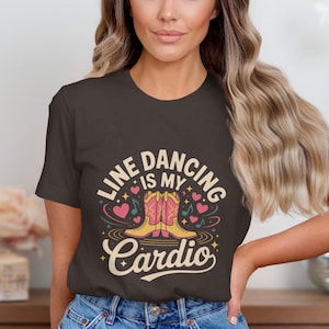 May include: A dark brown t-shirt with the text "LINE DANCING IS MY Cardio" in a retro design. The graphic includes cowboy boots, hearts, and musical notes. The shirt is worn by a person in blue jeans.