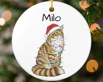May include: A round, white Christmas ornament featuring a tabby cat wearing a Santa hat and wrapped in colorful Christmas lights. The name "Milo" is printed above the cat in black letters. A gold string is attached for hanging.