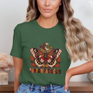 Op de afbeelding: Een donker olijfgroen t-shirt met korte mouwen en een centrale afbeelding van een mot met ingewikkelde patronen, een maansikkel en herfstbladeren. Onder de mot staat de tekst PUMPKIN PATCH CLUB in een veelkleurig, vet, rond lettertype.