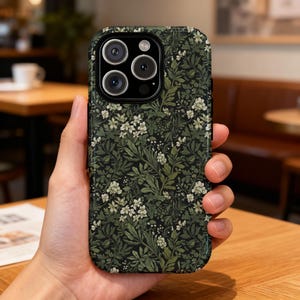 Op de afbeelding: Een telefoonhoesje met een donkergroen bloemenpatroon met witte bloemen en groene bladeren. De hoes heeft een zwarte rand en een camera-uitsparing met drie lenzen en een flitser.