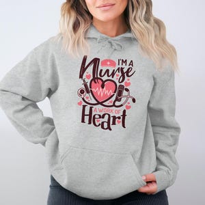 Op de afbeelding: Een lichtgrijze hoodie met een voorzak en een trekkoord. De voorkant van de hoodie heeft een afbeelding met de tekst "I'm a Nurse, A Work of Heart" met een hart, stethoscoop en medische symbolen in roze en kastanjebruin.