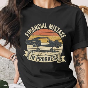 Op de afbeelding: Zwart T-shirt met een vintage afbeelding van een auto op krikken tegen een zonsondergang, met de tekst "FINANCIAL MISTAKE IN PROGRESS". Het ontwerp heeft een retro-stijl.