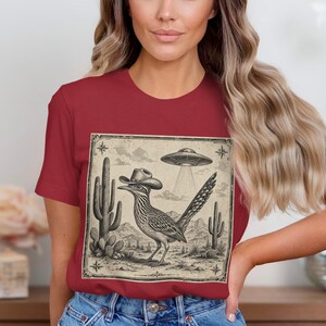 Op de afbeelding: Een bordeauxrood T-shirt met een vintage afbeelding. De afbeelding toont een roadrunner met een cowboyhoed, een UFO en een woestijnlandschap. De afbeelding is in zwart en wit.