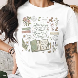 Op de afbeelding: Een wit t-shirt met een centrale afbeelding met de tekst Slow Down and Collect Beautiful Things. Het ontwerp bevat illustraties van een dagboek, een pen, gedroogde bloemen, bladeren, stempels en tape in gedempte groen-, roze- en bruintinten.