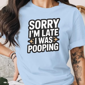 Op de afbeelding: Lichtblauw T-shirt met de tekst "SORRY I'M LATE I WAS POOPING" in zwart en wit. De tekst staat in het midden op de voorkant, met sterretjes. Het shirt heeft korte mouwen.