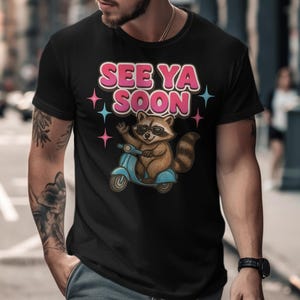 Op de afbeelding: Zwart T-shirt met een cartoon wasbeer met een zonnebril op en op een lichtblauwe scooter. De tekst "SEE YA SOON" staat boven de wasbeer in roze en witte letters, met steraccenten.