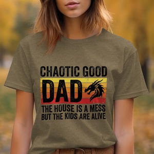 Op de afbeelding: Een olijfgroen t-shirt met een afbeelding op een retro zonsondergang-achtergrond in geel, oranje en rood. De tekst "CHAOTIC GOOD DAD" en "THE HOUSE IS A MESS BUT THE KIDS ARE ALIVE" staat in het zwart, vergezeld door een zwarte draak-silhouet.