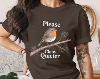 May include: A brown t-shirt with a detailed illustration of a robin on a branch with pine needles. The text "Please Chew Quieter" is above and below the bird. The shirt is a solid colour.