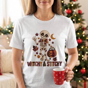 Op de afbeelding: Een wit T-shirt met korte mouwen en een afbeelding van een skelet met een geruite vlinderdas die een pompoenemmer vasthoudt. Het ontwerp bevat herfstbladeren, sterren, een maansikkel en de tekst "WITCHY & STITCHY" in een kleurrijk lettertype.