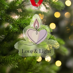 Puede incluir: Adorno navideño de acrílico transparente con diseño de vela. El adorno presenta una vela en forma de corazón con una llama, y las palabras "Love & Light" están impresas en la parte inferior. Cuelga de una cinta roja.