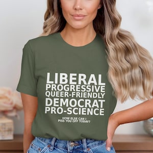 May include: An olive green t-shirt with white text: "LIBERAL PROGRESSIVE QUEER-FRIENDLY DEMOCRAT PRO-SCIENCE." The text "HOW ELSE CAN I PISS YOU OFF TODAY?" is below. The shirt is paired with blue jeans.