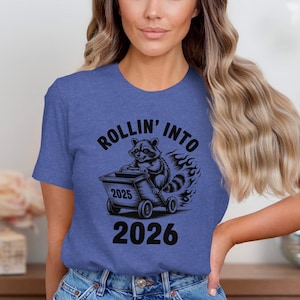 Puede incluir: Camiseta azul con un diseño de un mapache empujando un carro con los años 2025 y 2026. El texto "ROLLIN' INTO" está sobre el mapache. Las llamas sugieren movimiento.