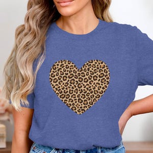 Op de afbeelding: Een blauw t-shirt met een groot hartvormig luipaardprintontwerp. De luipaardprint heeft een bruinachtige achtergrond met donkerbruine vlekken. Het shirt heeft korte mouwen en een ronde hals.