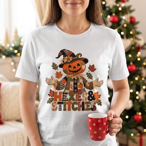 Op de afbeelding: Een wit t-shirt met korte mouwen en een afbeelding van een vogelverschrikker met een pompoenhoofd, gekleed in een hoed en kleding van patchwork. Het ontwerp bevat herfstbladeren en de tekst "HEXES & STITCHES" in een patchwork-stijl lettertype.