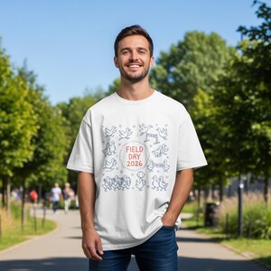 Op de afbeelding: Een wit t-shirt met blauwe eendenillustraties en de tekst "FIELD DAY 2026" in een rode cirkel. De persoon draagt het t-shirt buiten op een zonnige dag, met bomen op de achtergrond.